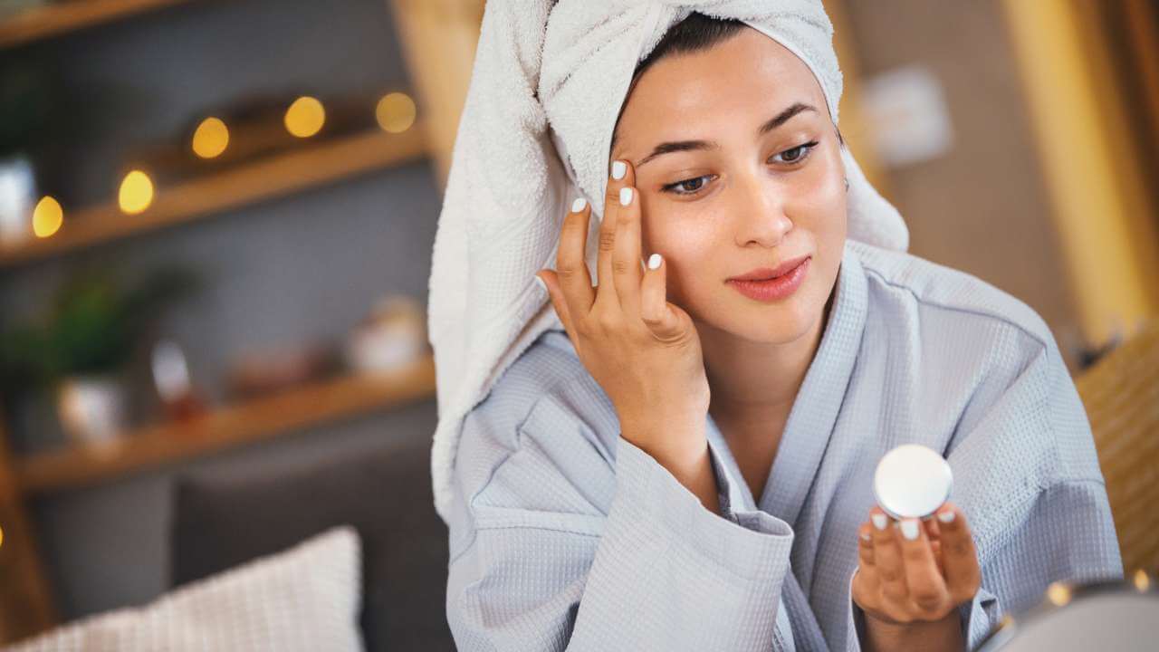Woman in robe with hair wrapped in a towel applies a cosmetic skin whitening agent for sun spots to her face.