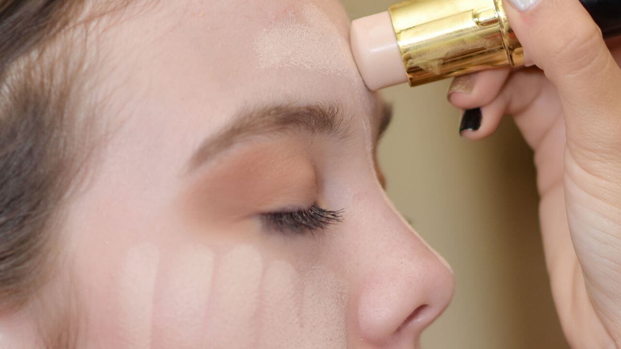 Closeup, right-sided view of woman’s face from nose to hairline with a liquid foundation stick being applied to her forehead.