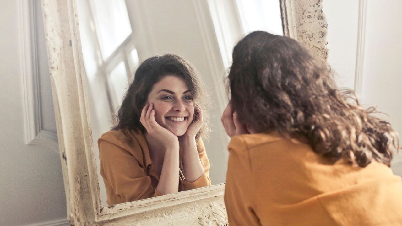Happy woman smiling at her reflection in mirror with elbows propped up on dresser while supporting her chin in both hands.