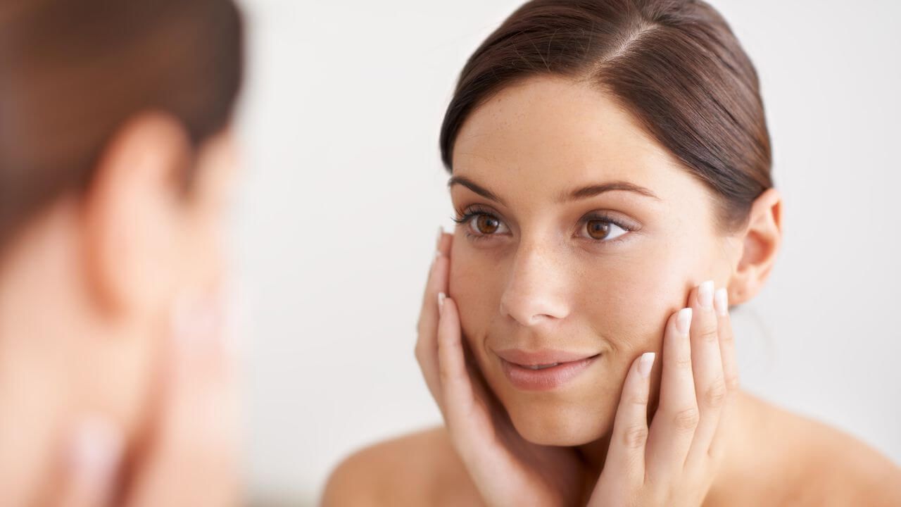 Young woman is pleased with kojic acid's lightening effect while looking into a mirror and touching both sides of her face.