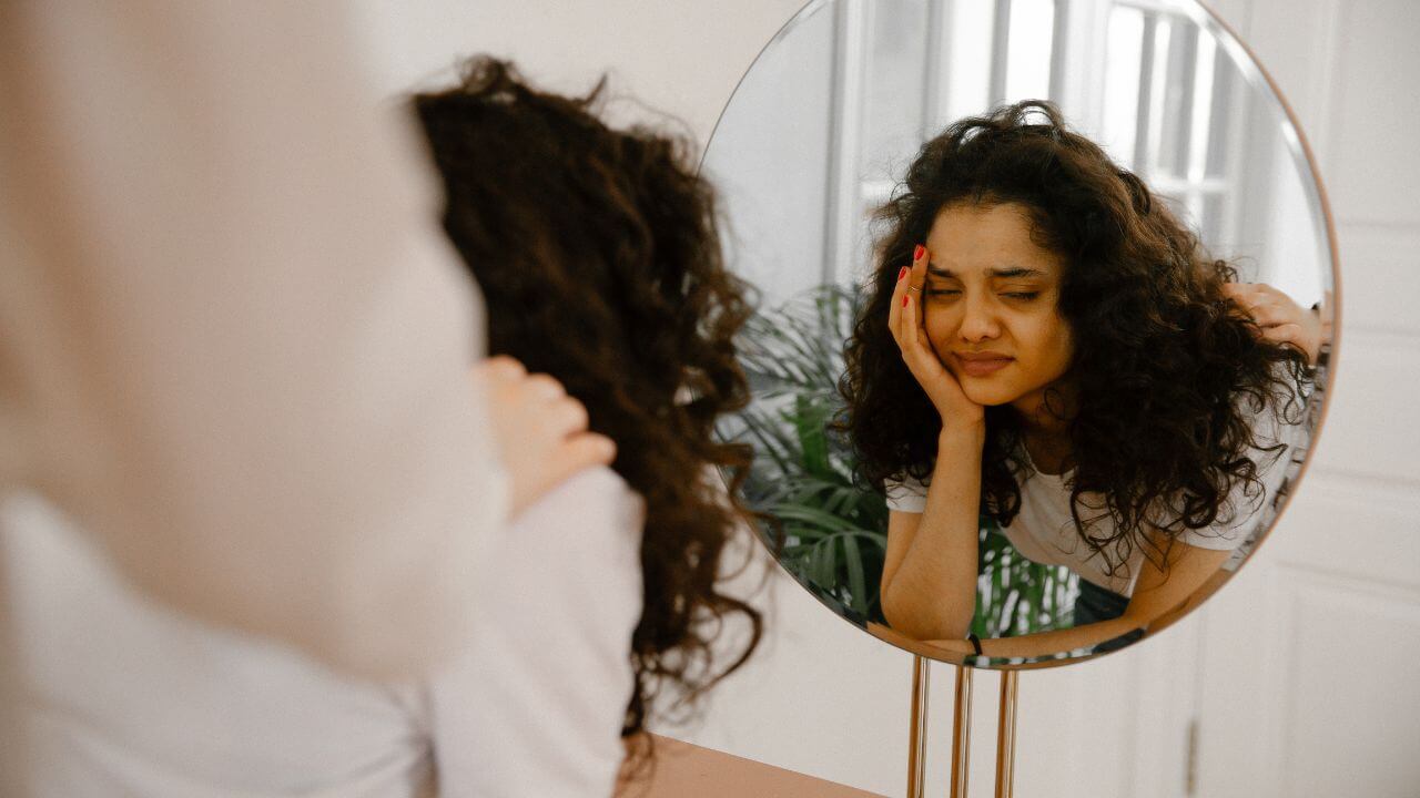 Reflection of sad woman in mirror with eyes closed, leaning over makeup table with another person’s hand on her shoulder.