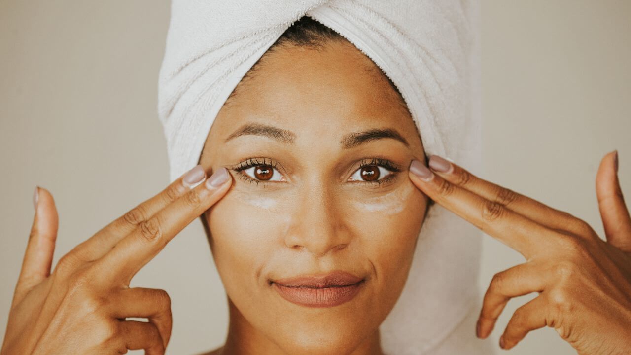 Close up of a smiling woman with hair wrapped in a towel, applying primer under both eyes with her index & middle fingers.