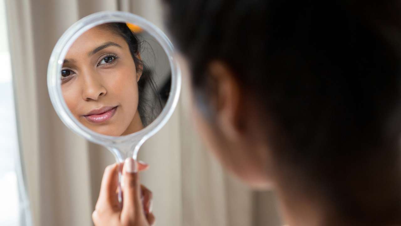 Reflection of a woman’s face in handheld mirror while she looks for different types of hyperpigmentation on her face.